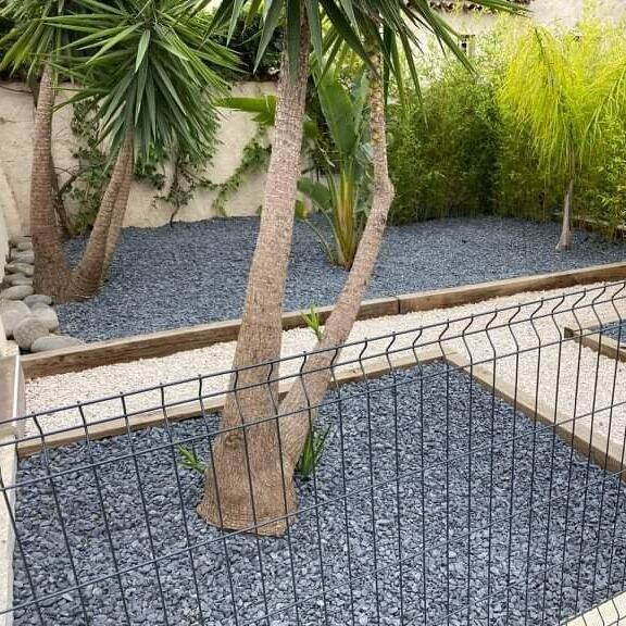Aménagement de Jardin Moderne avec Gravier Gris et Blanc Jardin moderne avec gravier gris et blanc, palmiers exotiques et clôture noire sur fond de bambous.