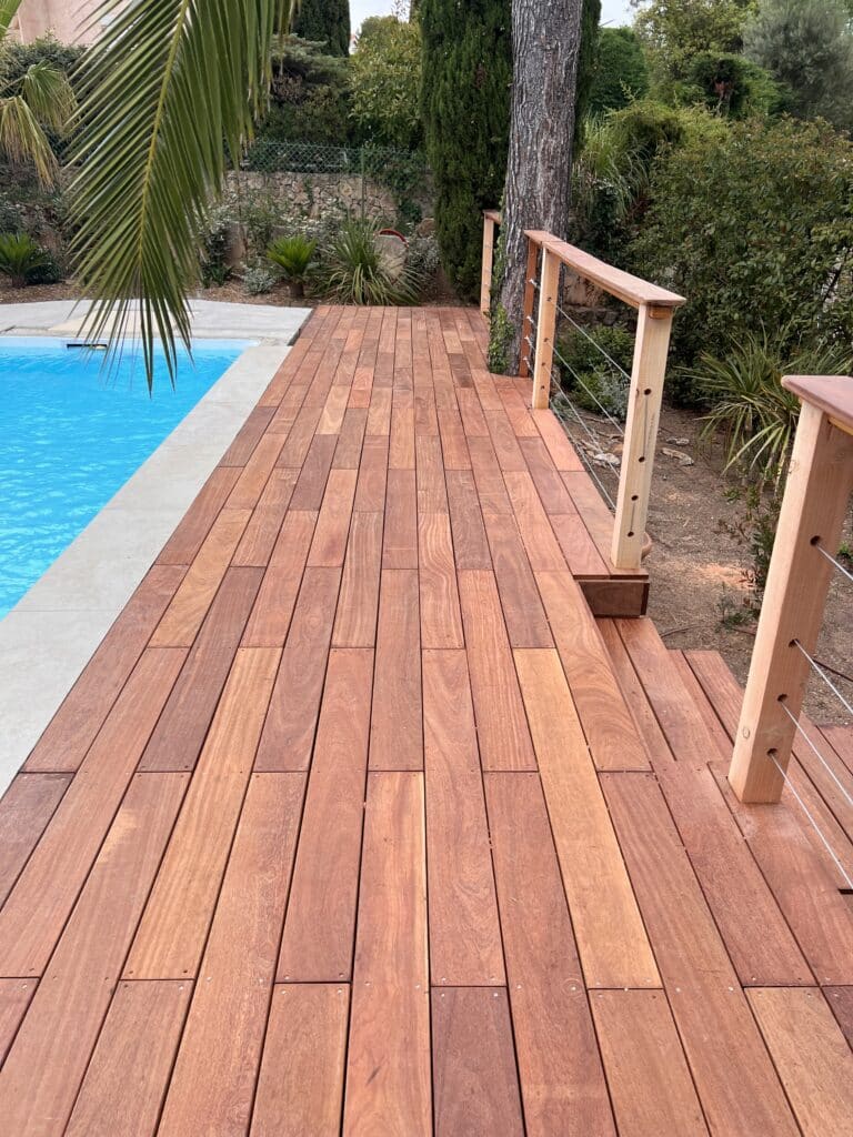 Terrasse en bois exotique pour piscine : aménagement chic Terrasse en bois exotique longeant une piscine bleue avec garde-corps en bois et câbles inox dans un jardin arboré.